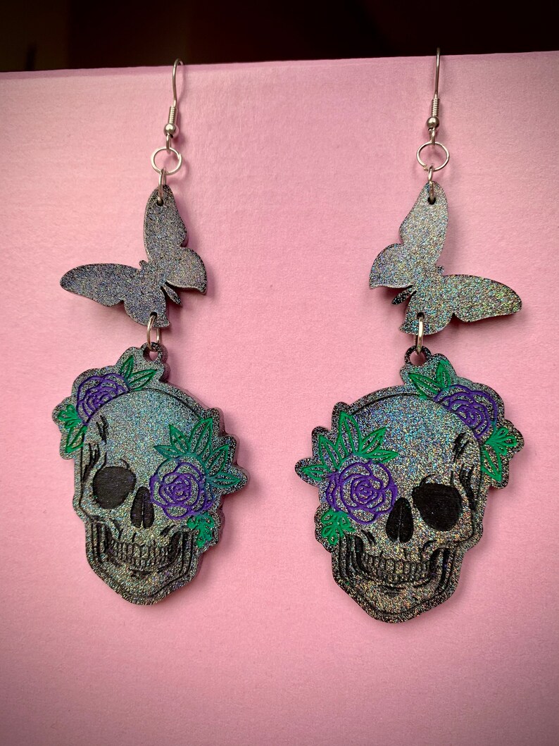May include: A pair of silver dangle earrings with a black glitter skull with purple and green floral accents and a black glitter butterfly charm.