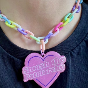 May include: A pastel-colored chain necklace with a heart-shaped pendant. The pendant is light pink with a dark pink border and the words "Smash the Patriarchy" in white. The chain links are in various pastel colors.