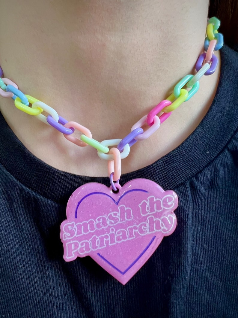 May include: A pastel-colored chain necklace with a heart-shaped pendant. The pendant is light pink with a dark pink border and the words "Smash the Patriarchy" in white. The chain links are in various pastel colors.