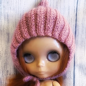 May include: A pink knitted doll hat with a pointed top. The hat has a ribbed texture and is worn by a doll with large, white eyes.