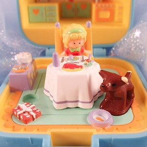 Puede incluir: Escena de casa de muñecas en miniatura con una muñeca rubia sentada a la mesa. También hay una figura de perro marrón y una pequeña caja de regalo. La casa de muñecas es azul y amarilla.