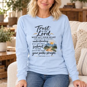 Op de afbeelding: Een lichtblauw shirt met lange mouwen en een grafische print met de tekst Trust in the Lord with all your heart and lean not on your own understanding; in all your ways submit to him, and he will make your paths straight. Proverbs 3:5-6.