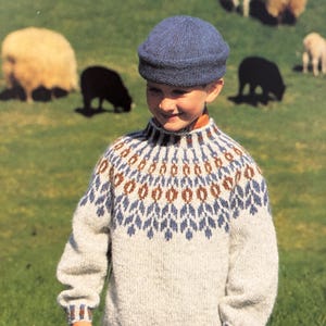 Könnte beinhalten: Ein kleiner Junge trägt einen weißen Pullover mit einem blauen und braunen gemusterten Joch, eine blaue Strickmütze und eine blaue Jeans. Er steht auf einem Feld mit Schafen im Hintergrund.
