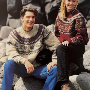 Könnte beinhalten: Zwei Personen tragen traditionelle isländische Pullover, bekannt als Lopapeysa, mit komplizierten Mustern in Braun-, Weiß- und Blautönen. Sie sitzen im Freien auf Felsen.