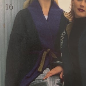 Può includere: Un cappotto grigio scuro con colletto blu scuro e cintura abbinata. Il cappotto è indossato sopra una gonna argentata a righe verticali. Il numero 16 è visibile in alto a sinistra.