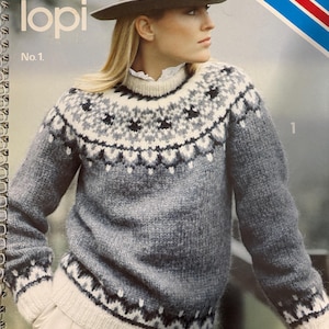 Puede incluir: Una mujer con un suéter gris con canesú y puños estampados, y un sombrero gris. El suéter es de Alafoss lopi, No. 1. El suéter tiene un diseño blanco y negro alrededor del cuello y los puños.