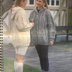 Op de afbeelding: Twee vrouwen in kabelgebreide capuchontruien. De ene is crèmekleurig, de andere grijs. De vrouw links draagt een witte short en kniehoge sokken. De vrouw rechts draagt een zwarte broek en grijze sokken. Afbeelding uit een catalogus.