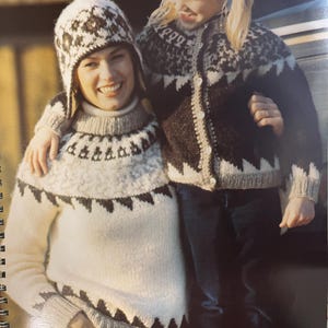 Puede incluir: Una mujer y un niño con jerséis y gorros de punto a juego. Los jerséis son blancos con diseños geométricos negros y grises. El gorro tiene orejeras y un diseño a juego. El niño lleva una rebeca abotonada con el mismo diseño.