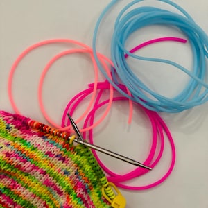 Peut inclure: Gros plan sur un projet de tricotage coloré avec un mélange de fils roses, jaunes, verts et bleus. Des aiguilles à tricoter argentées sont insérées dans le tissu tricoté, avec un marqueur de point jaune. Plusieurs aiguilles à tricoter circulaires roses et bleues sont également visibles.