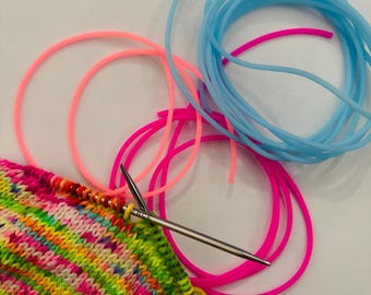 Könnte beinhalten: Nahaufnahme eines Strickprojekts mit einer bunten Mischung aus rosa, gelber, grüner und blauer Wolle. Silberne Stricknadeln sind in den gestrickten Stoff eingeführt, mit einem gelben Maschenmarkierer. Mehrere Rundstricknadeln in Rosa und Blau sind ebenfalls sichtbar.