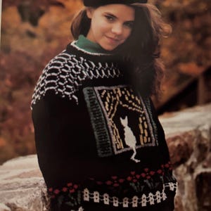 May include: A black sweater with a white cat design and a green, white and yellow border. The sweater has a red, white and green floral pattern at the bottom.