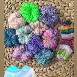 May include: A collection of ten small, round yarn bundles in various colours including neon green, pastel pink, purple, and variegated blue and pink tones, accompanied by a single knitted sock with horizontal stripes in rainbow colours.