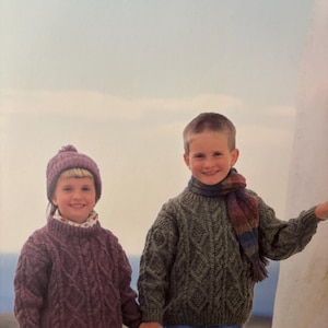 Può includere: Due bambini che indossano maglioni e cappelli di lana. La ragazza indossa un maglione viola e un cappello bianco con un pon pon. Il ragazzo indossa un maglione verde e una sciarpa a righe.