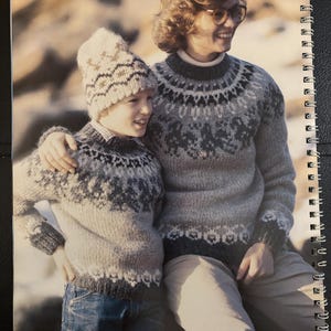 May include: Two people wearing matching gray and white patterned sweaters and a hat. The sweaters have a round yoke design with a repeating pattern. The hat has a similar design. The text "9-10-11" is visible at the top of the image, and "Alafoss lopi" is at the bottom.