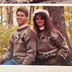 May include: A couple wearing matching brown knit sweaters with duck designs. The sweaters have a crew neck and feature a pattern of ducks in the center. The background is a blurred forest scene with fall colors.