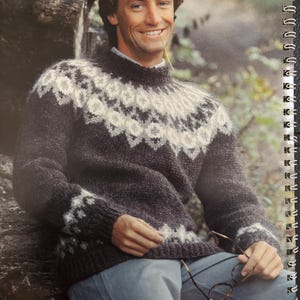May include: A dark grey and white patterned sweater with a high neckline. The sweater has a geometric design around the yoke and cuffs. The image also shows a person wearing the sweater, holding glasses.