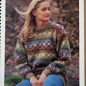 Può includere: Una donna indossa un maglione a collo alto con motivi marroni, verdi, blu e bianchi. Il maglione ha un design geometrico. La donna è seduta e indossa jeans blu. Il testo "Available in Kit Form" è nell'angolo in alto a destra dell'immagine.