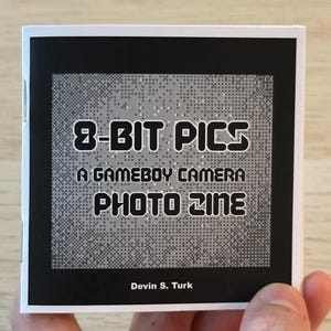 以下が含まれることがあります： 「8-BIT PICS A GAMEBOY CAMERA PHOTO ZINE」というタイトルの小さな正方形のジン。白文字が黒い背景に書かれています。カバーはピクセル化されたデザインです。著者のDevin S. Turkの名前が下部に印刷されています。ジンは手で持たれています。