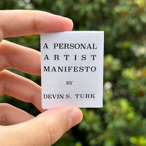 Op de afbeelding: Een klein wit kaartje met zwarte tekst waarop staat "A Personal Artist Manifesto by Devin S. Turk".