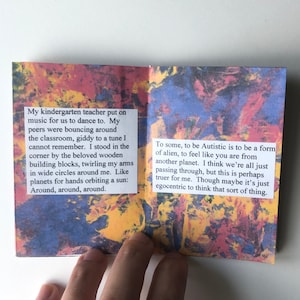 Puede incluir: Un peque&ntilde;o libro colorido con una p&aacute;gina blanca que muestra texto sobre un maestro de jard&iacute;n de infantes que pone m&uacute;sica para que la clase baile. El texto de la p&aacute;gina derecha habla sobre la sensaci&oacute;n de ser autista y lo compara con ser de otro planeta. El libro tiene un fondo abstracto colorido.