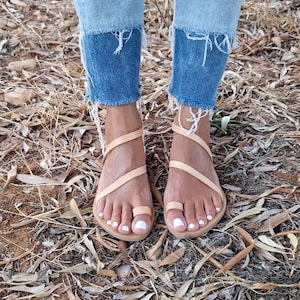 K&ouml;nnte beinhalten: Ein Paar hellbraune Ledersandalen mit mehreren Riemen, die sich um die Kn&ouml;chel und Zehen wickeln. Die Sandalen werden von einer Person mit nackten F&uuml;&szlig;en und wei&szlig;em Nagellack getragen.