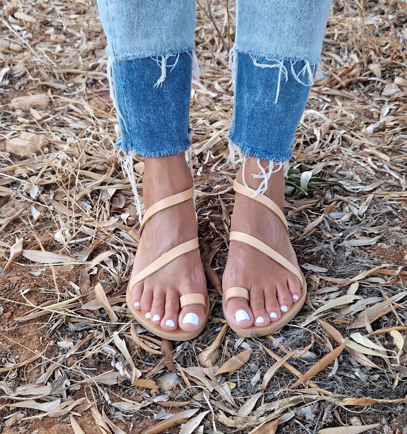 K&ouml;nnte beinhalten: Ein Paar hellbraune Ledersandalen mit mehreren Riemen, die sich um die Kn&ouml;chel und Zehen wickeln. Die Sandalen werden von einer Person mit nackten F&uuml;&szlig;en und wei&szlig;em Nagellack getragen.
