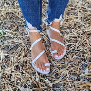 K&ouml;nnte beinhalten: Wei&szlig;e Ledersandalen mit mehreren Riemen, die sich um Fu&szlig; und Kn&ouml;chel wickeln. Die Sandalen werden von einer Person getragen, die auf trockenem Gras steht.