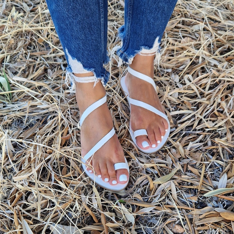 K&ouml;nnte beinhalten: Wei&szlig;e Ledersandalen mit mehreren Riemen, die sich um Fu&szlig; und Kn&ouml;chel wickeln. Die Sandalen werden von einer Person getragen, die auf trockenem Gras steht.