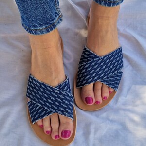 K&ouml;nnte beinhalten: Ein Paar blaue Jeans-Sandalen mit gekreuzten Riemen. Die Sandalen haben eine braune Ledersohle und werden an den F&uuml;&szlig;en getragen.