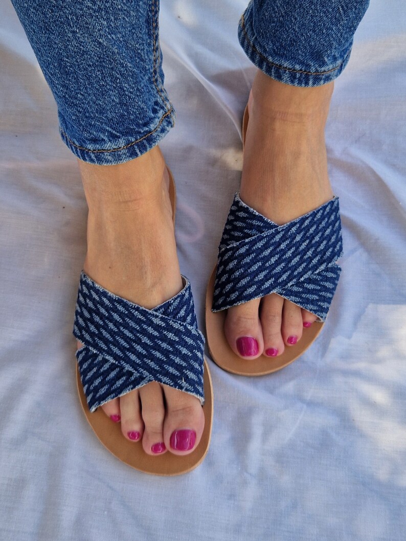 K&ouml;nnte beinhalten: Ein Paar blaue Jeans-Sandalen mit gekreuzten Riemen. Die Sandalen haben eine braune Ledersohle und werden an den F&uuml;&szlig;en getragen.