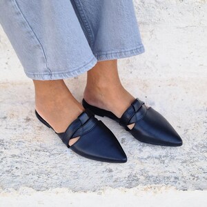 May include: A pair of black leather pointed-toe mules with a braided strap detail. The mules are worn on a person's feet.