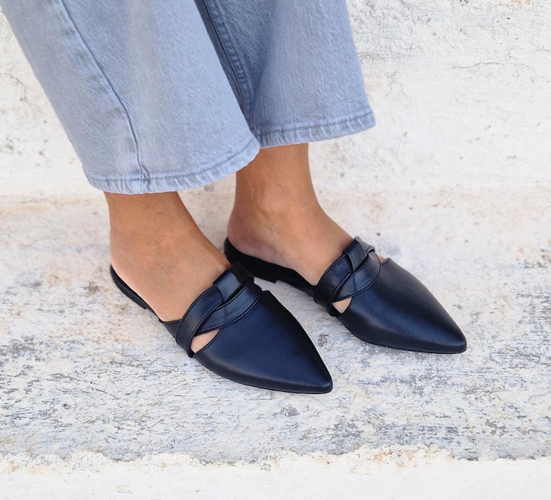 May include: A pair of black leather pointed-toe mules with a braided strap detail. The mules are worn on a person's feet.