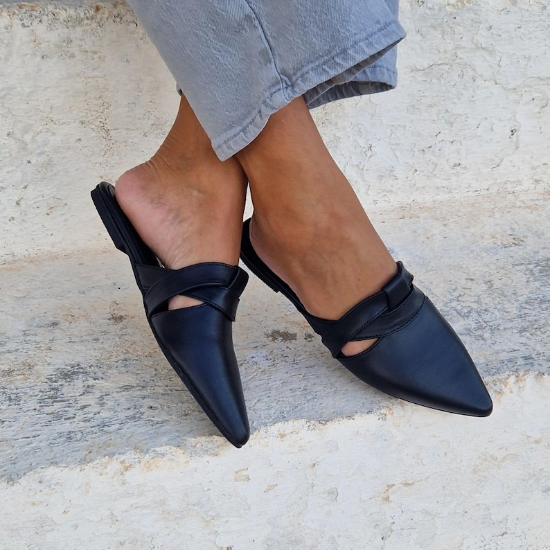 May include: A pair of black leather pointed-toe mules with a criss-cross strap detail. The mules are worn on a person's feet.