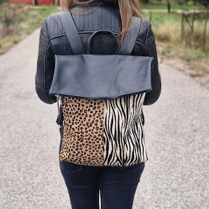 Puede incluir: Mochila de cuero negro con cierre de solapa. La parte inferior de la bolsa presenta un estampado de leopardo y cebra. Las correas y el asa superior son negras.