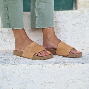 以下が含まれることがあります： 厚底のタンレザーのフラットサンダル。裸足にオレンジ色のマニキュアを塗った人が履いています。