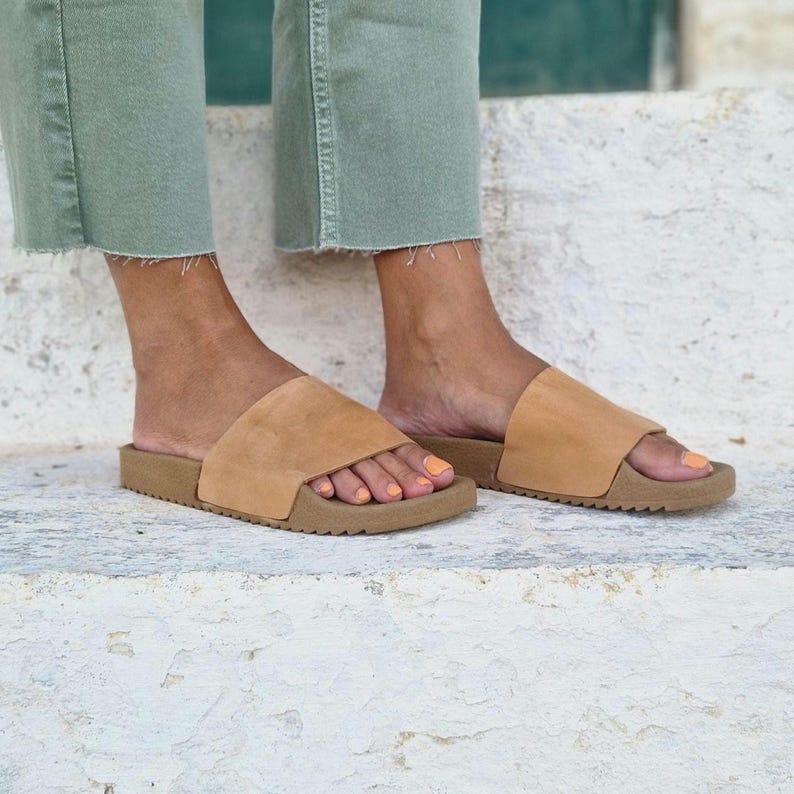 以下が含まれることがあります： 厚底のタンレザーのフラットサンダル。裸足にオレンジ色のマニキュアを塗った人が履いています。