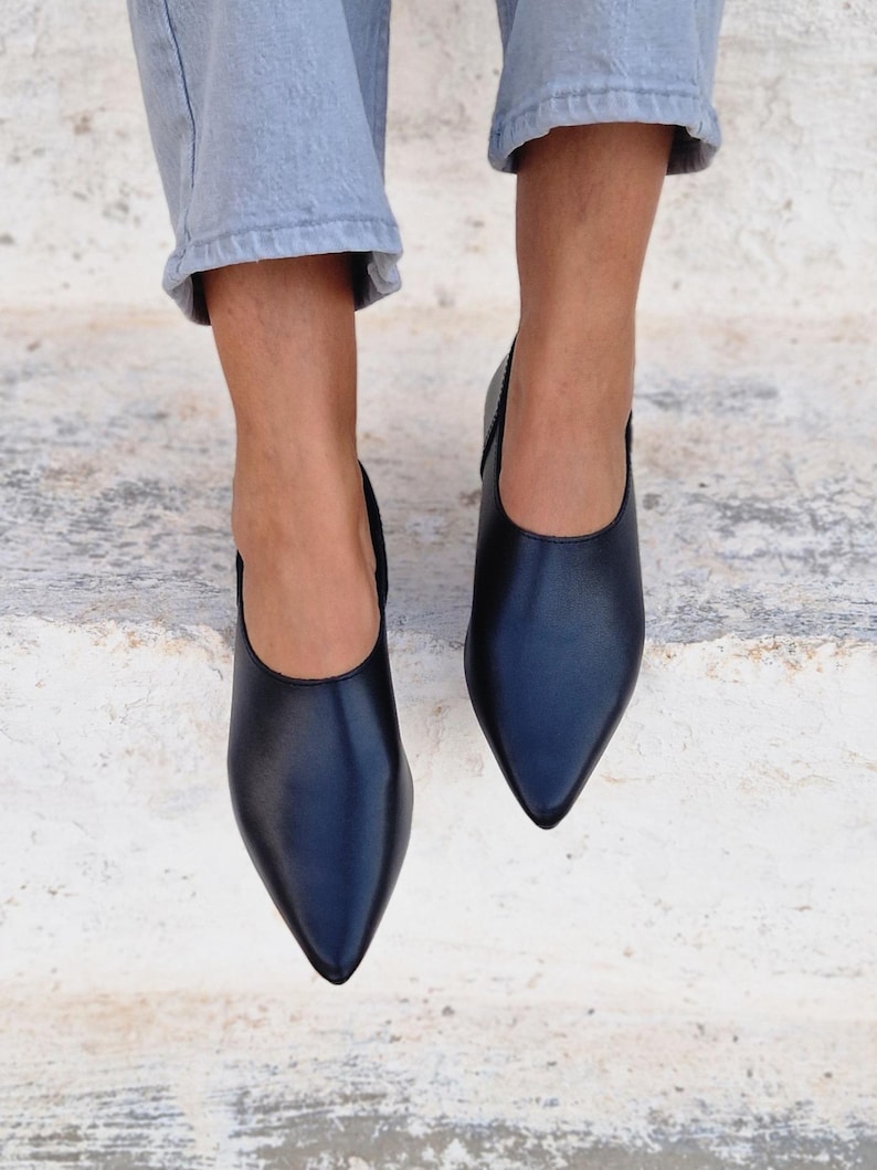 Pode incluir: Um par de sapatos pretos de couro de bico fino com salto baixo. Os sapatos s&atilde;o usados por uma pessoa que est&aacute; vestindo jeans azul claro.
