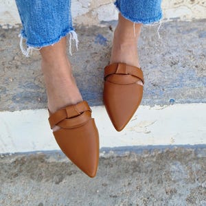 May include: A pair of brown leather pointed toe mules with a knotted strap detail. The mules are worn on a person's feet with denim jeans.
