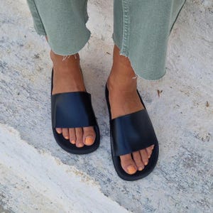 以下が含まれることがあります： シンプルなデザインの黒い革製のサンダル。フラットなソールと足の甲に渡る幅広のストラップが特徴です。