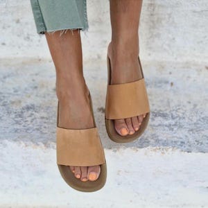 以下が含まれることがあります： シンプルなデザインのタンレザーのフラットサンダル。サンダルはフラットソールで、足の甲に幅広のストラップが付いています。