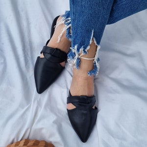 May include: A pair of black leather pointed-toe mules with a cut-out design and a knotted detail on the strap. The mules are worn on a person's feet.