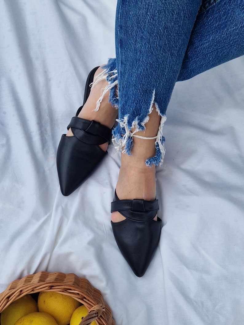 May include: A pair of black leather pointed-toe mules with a cut-out design and a knotted detail on the strap. The mules are worn on a person's feet.