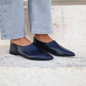 Pode incluir: Um par de sapatos pretos de couro sem cadar&ccedil;os com bico fino. Os sapatos s&atilde;o planos e t&ecirc;m um design simples.