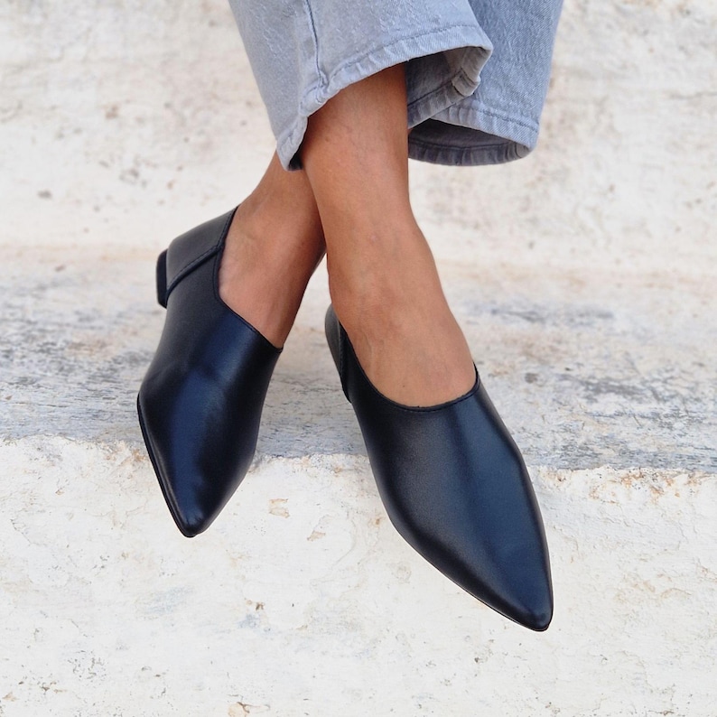 Pode incluir: Um par de sapatos baixos de bico preto de couro com um design de deslizamento. Os sapatos s&atilde;o usados por uma pessoa sentada em um degrau de pedra branca.