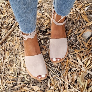 Peut inclure: Une paire de sandales à bride cheville beige clair avec un bout pointu. Les sandales sont en daim et ont une semelle plate. Les sandales sont portées par une personne debout sur un lit de feuilles sèches.
