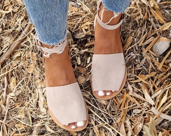 May include: A pair of light beige ankle strap sandals with a pointed toe. The sandals are made of suede and have a flat sole. The sandals are worn by a person standing on a bed of dry leaves.
