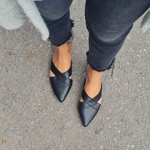 May include: A pair of black leather pointed-toe flats with criss-cross straps. The shoes are worn by a person wearing black jeans.