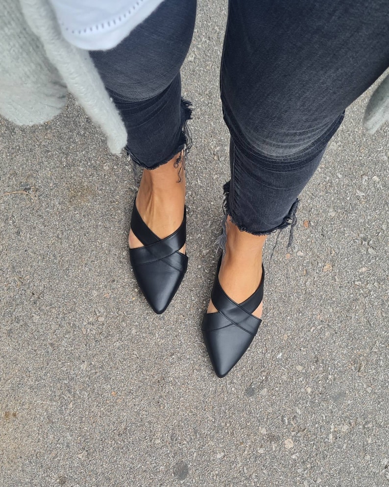 May include: A pair of black leather pointed-toe flats with criss-cross straps. The shoes are worn by a person wearing black jeans.