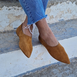 Pode incluir: Um par de sapatos baixos de camur&ccedil;a marrom com bico fino. Os sapatos s&atilde;o usados por uma pessoa que est&aacute; vestindo jeans azul.
