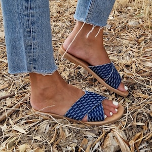 K&ouml;nnte beinhalten: Ein Paar blaue Jeans-Sandalen mit einem gekreuzten Riemen-Design. Die Sandalen haben eine braune Ledersohle und werden an den F&uuml;&szlig;en getragen.